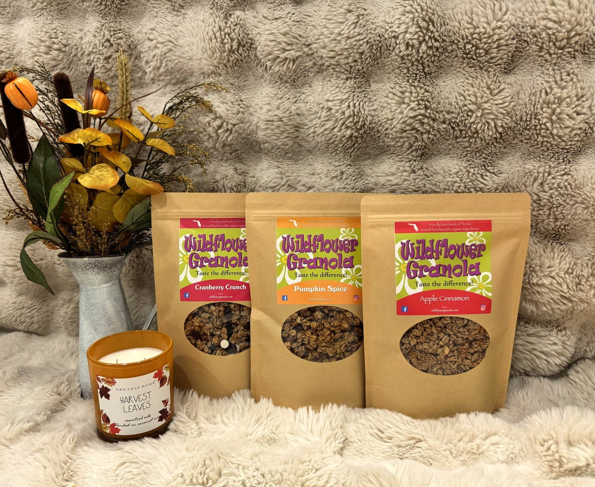 Apple Cinnamon, Pumpkin Spice and Cranberry Crunch granola bags on a cream blanket with a fall floral arrangement and a lit candle